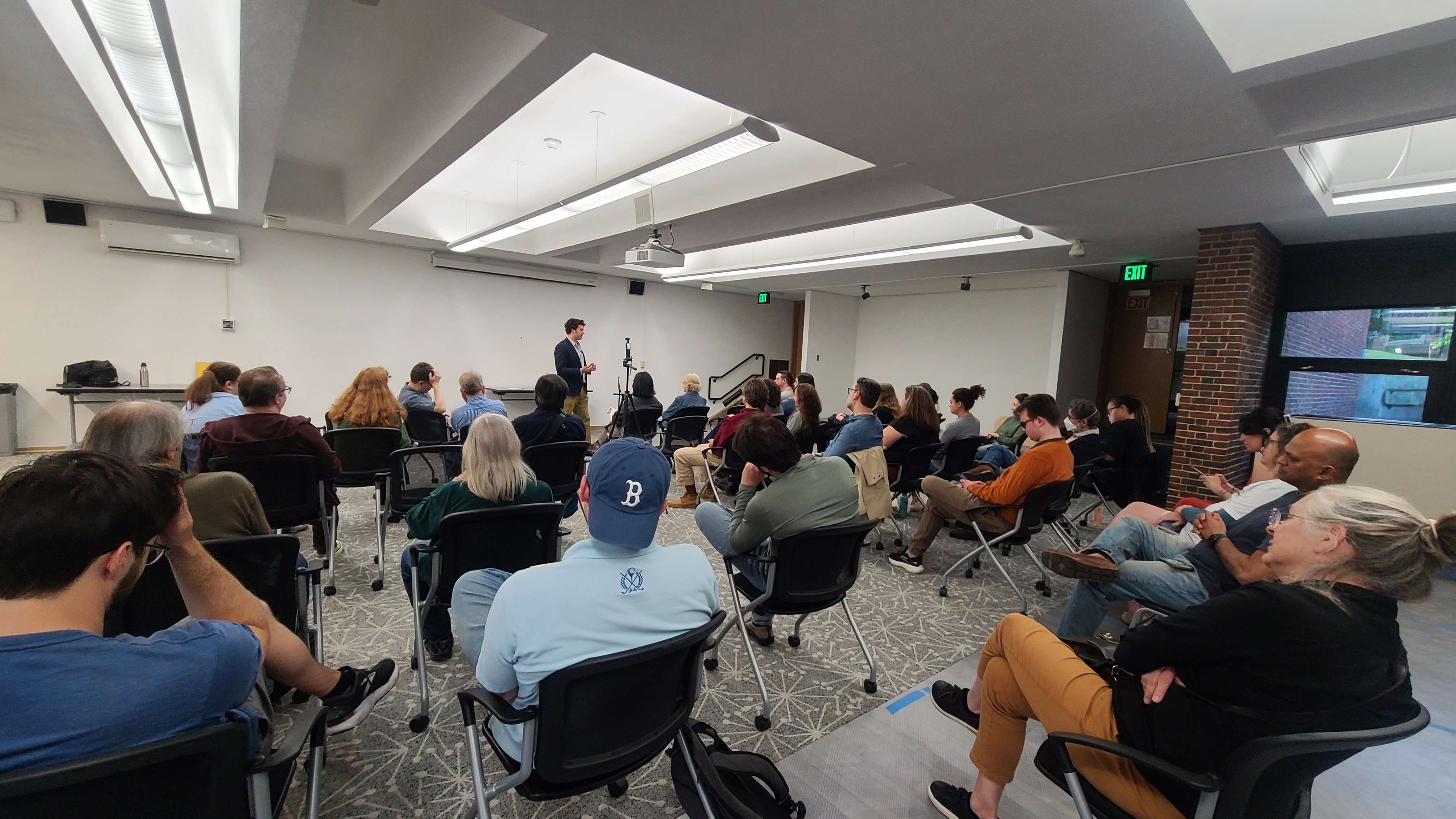 Jason Poulos addressing a full room of seated attendees at the Brookline town hall, Coolidge Corner Library, June 2, 2026